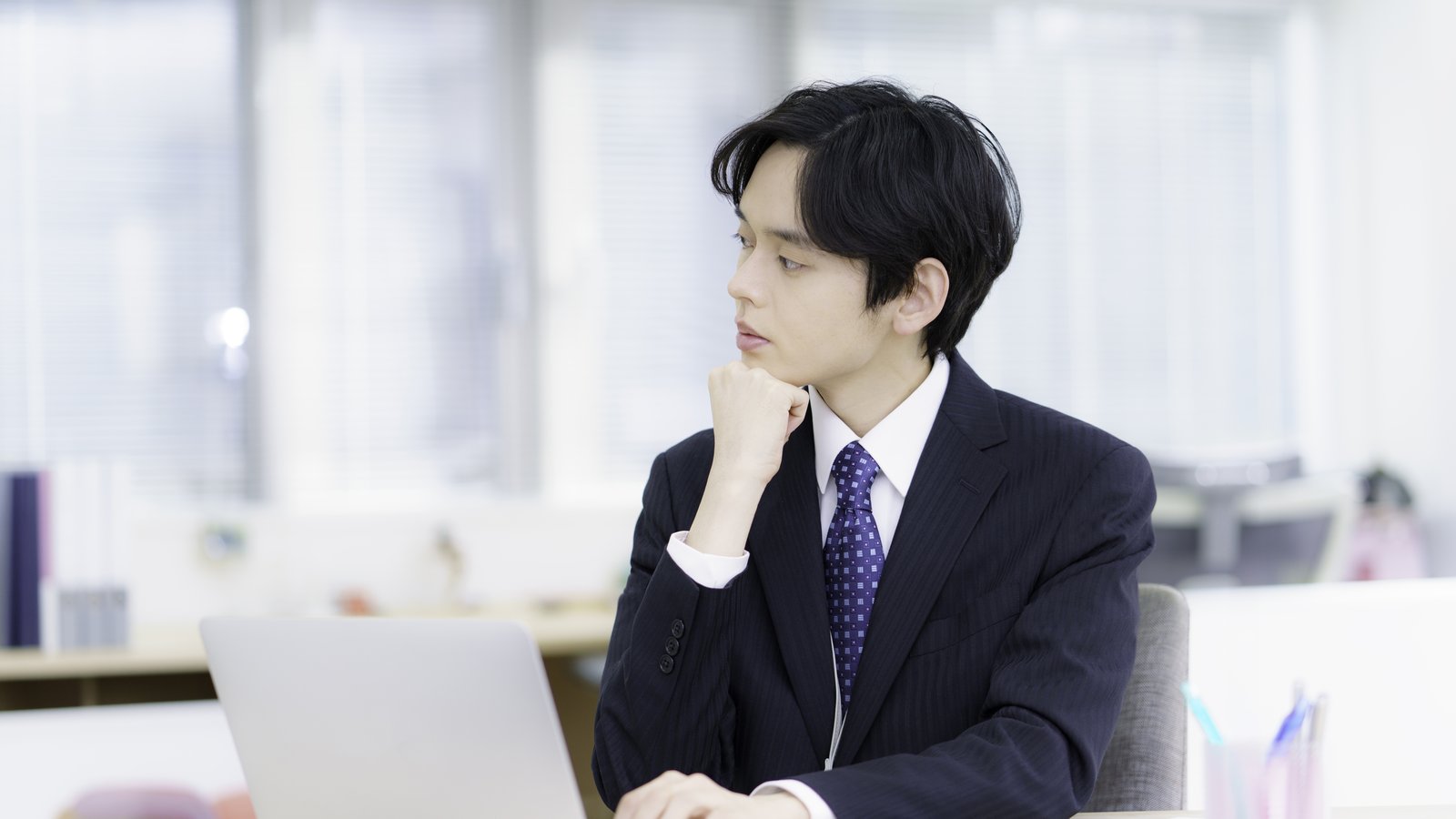 オフィスでノートPCを前に考え込むスーツ姿の日本人男性